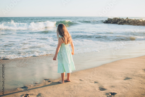 girl in a blue dress standing in the water waves at the beach with her back to the camera