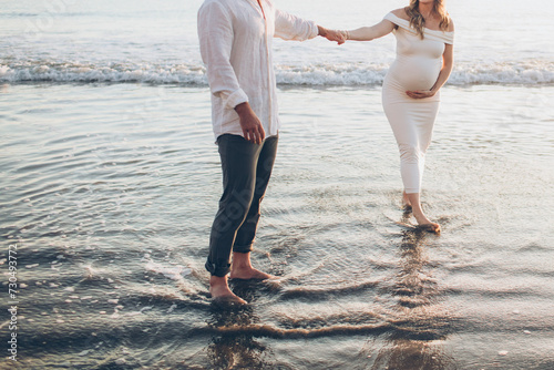 close up crop photo of a pregnant maternity couple holding hands looking at each other standing in the water at sunset on the beach