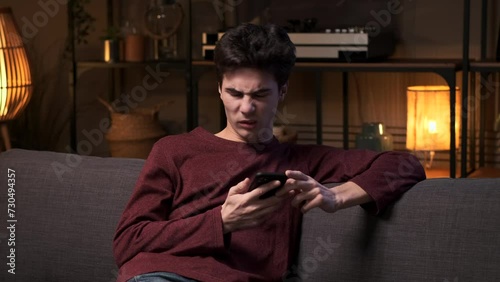 Caucasian man is using phone on the living room couch, sees bad news and reacts to it with disgust. Shock and obscene content, social media problems concept. photo