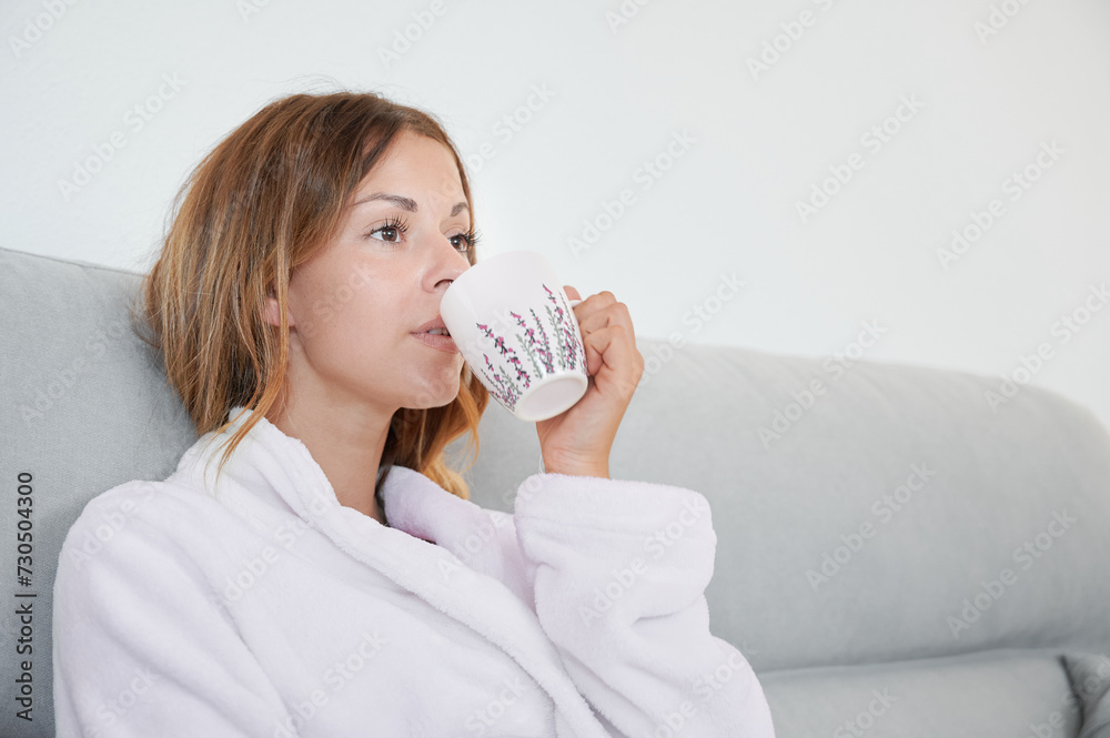 Young sleepy woman wrapped in a bathrobe drinks morning coffee while resting on the sofa. Typical home scenes from the weekend. Young sleepy woman wrapped in a bathrobe drinks morning coffee while resting on the sofa. Typical home scenes from the weekend.