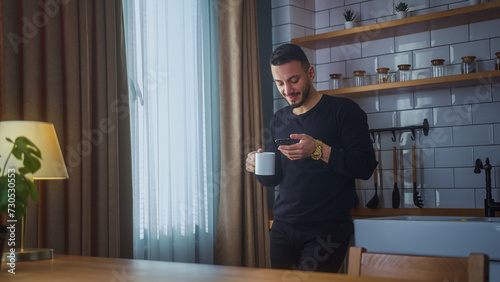 Smiling man standing in modern kitchen having morning coffee, holding cell phone scrolling news feed, checking emails, social media on smartphone