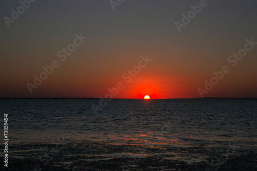 Sunset over the sea against the background of the coast. Evening on the sea calm in clear weather. The disk of the sun in the evening sky over the sea. © Vladimir