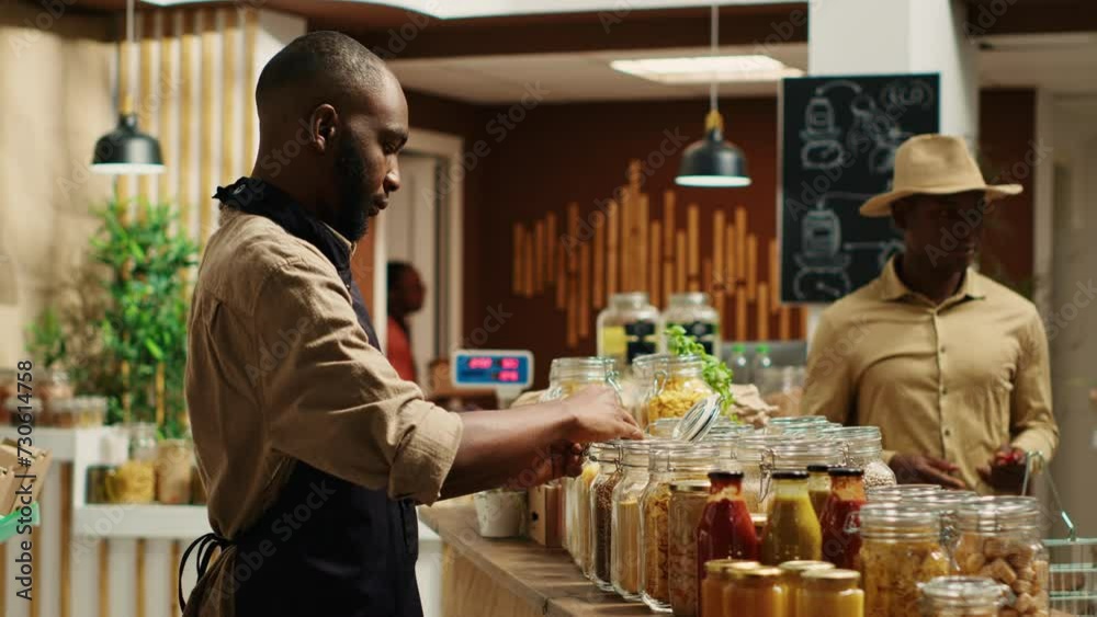 African american farmer opening local zero waste store with natural additives free bulk products and homegrown produce. Merchant preparing goods in reusable jars at eco supermarket. Camera A. African american farmer opening local zero waste store with natural additives free bulk products and homegrown produce. Merchant preparing goods in reusable jars at eco supermarket. Camera A.