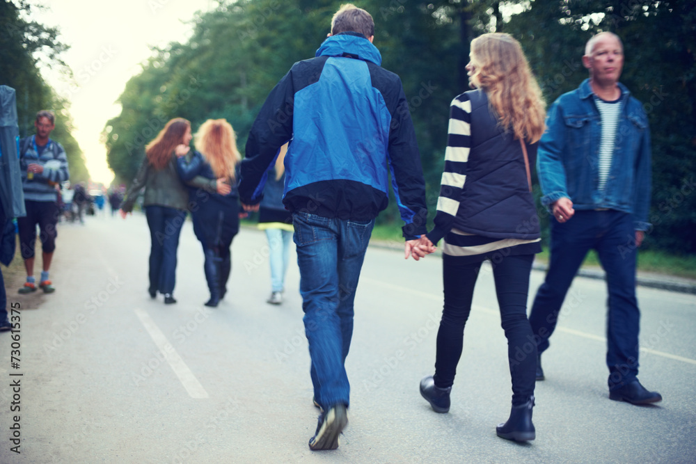 Obraz premium Couple, back and holding hands while walking among trees and crowds along road in New Zealand. Rear view of man, woman and people for travel, date and bonding in busy woods, park and highway