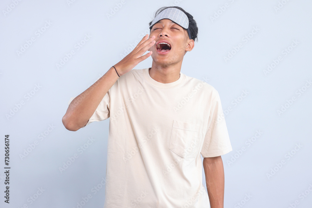 Tired Asian guy with sleep mask standing and yawning Tired Asian guy with sleep mask standing and yawning
