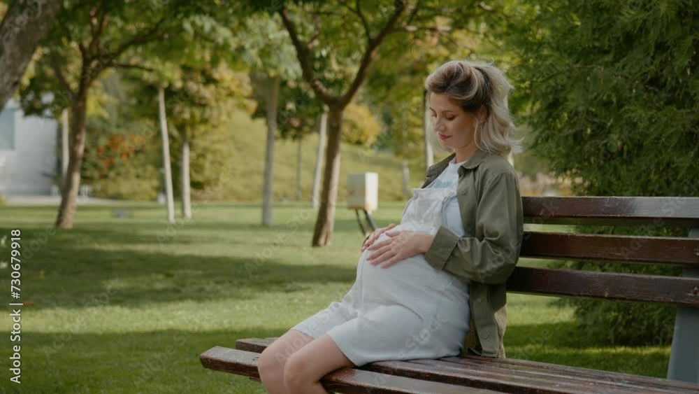 Calm pregnant female sitting on park bench holds bump surrounded by nature outdoors. Female enjoying maternity. Carrying a baby, maternity concept. Calm pregnant female sitting on park bench holds bump surrounded by nature outdoors. Female enjoying maternity. Carrying a baby, maternity concept.