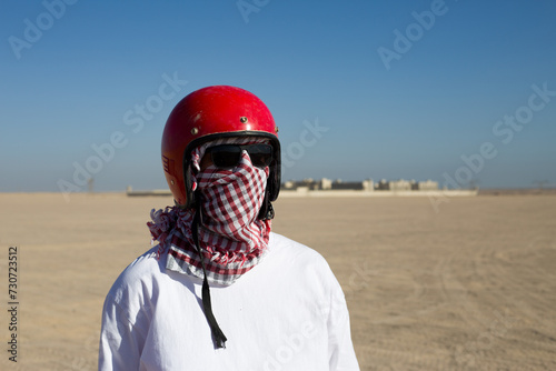 Man wearing keffiyeh and a helmet wrapped around his face stands against the backdrop of desert during a safari photo