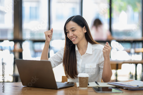 Fotografie A delighted Asian businesswoman raises her fists in victory while looking at her laptop in a well-lit office space