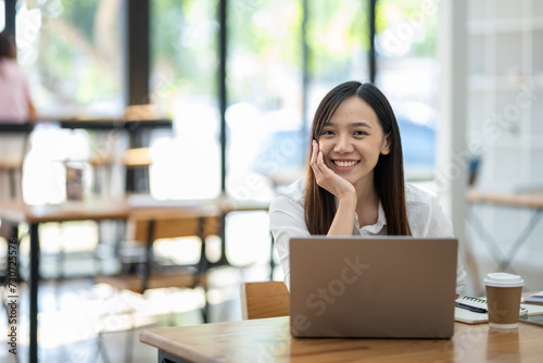 A smiling Asian businesswoman enjoying her work on a laptop, comfortably seated at a wooden table in a casual office setting..