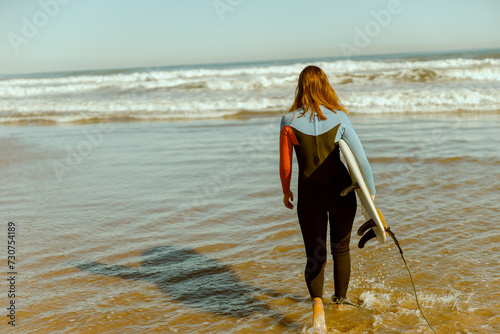 Back view of female surfer in wetsuit with his surfboard entering the sea for riding on waves