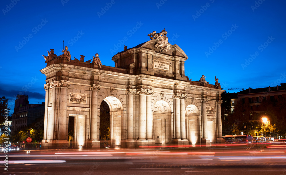 Fototapeta premium Puerta de Alcalá by the entrance of El Retiro park at blue hour in Madrid. Puerta Alcala evening view with car light trails as the night approaches. Downtown vibe on the Spanish capital busy streets