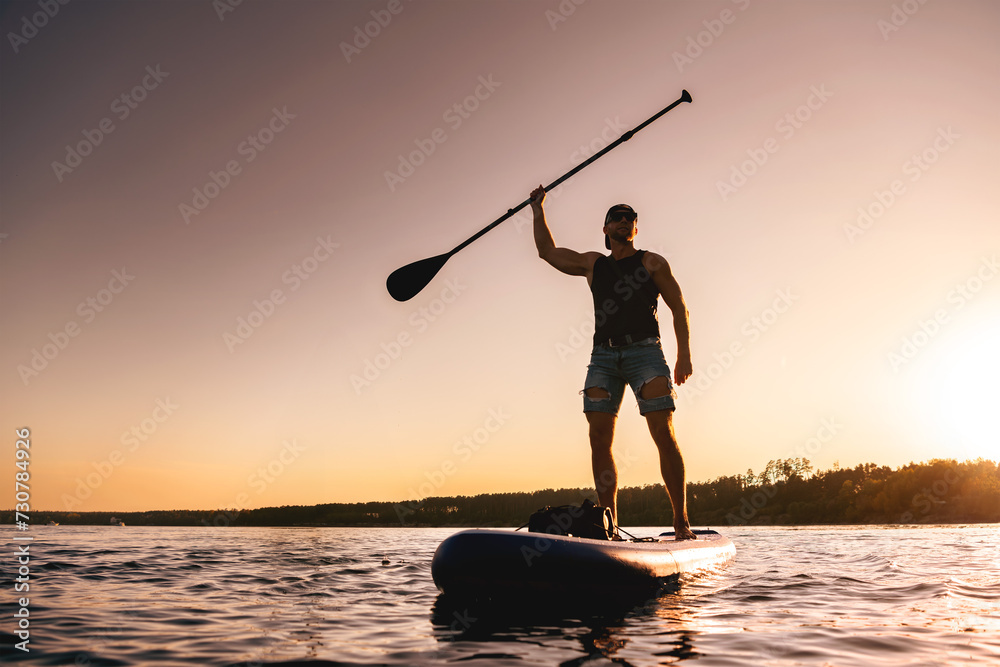 Happy sporty surfer is standing on sup board with paddle in hand at sunset lake