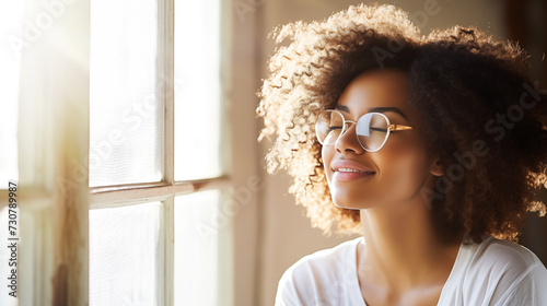 Woman in white t-shirt and glasses, standing by the window with gentle sunlight casting a warm glow, eyes closed and a subtle smile, embodying serene joy photo