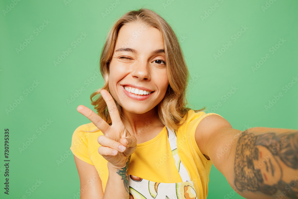 Close up young housewife housekeeper chef cook baker woman she wears apron yellow t-shirt do selfie shot on mobile cell phone show v-sign wink isolated on plain green background. Cooking food concept. Close up young housewife housekeeper chef cook baker woman she wears apron yellow t-shirt do selfie shot on mobile cell phone show v-sign wink isolated on plain green background. Cooking food concept.
