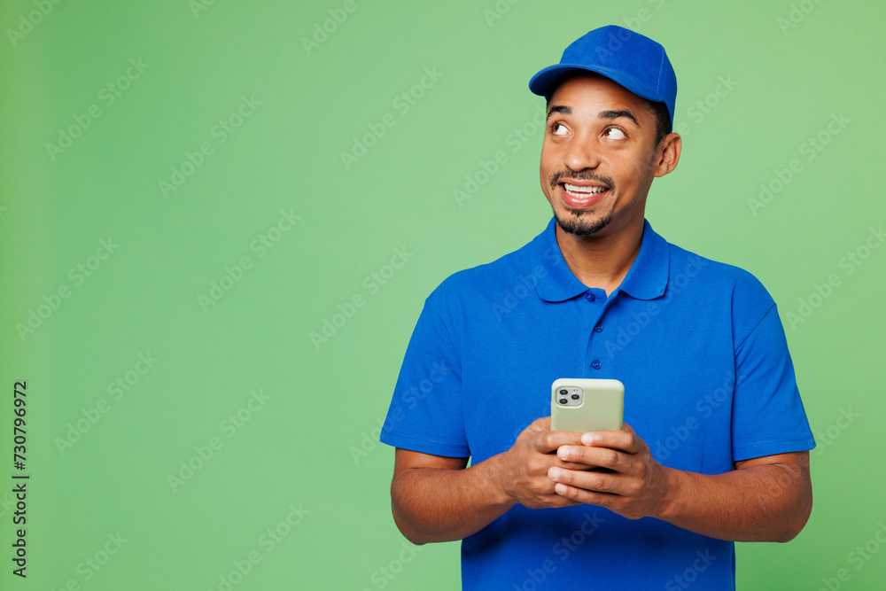 Professional delivery guy employee man wear blue cap t-shirt uniform workwear work as dealer courier hold use mobile cell phone look aside on area isolated on plain green background. Service concept. Professional delivery guy employee man wear blue cap t-shirt uniform workwear work as dealer courier hold use mobile cell phone look aside on area isolated on plain green background. Service concept.