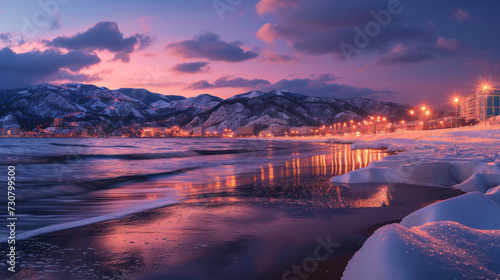 View of sandy beach, sea, waves, snowy mountains, street lights at winter night. Landscape with ocean, reflection, illumination, snowy rocks in clouds, pink sky at sunset