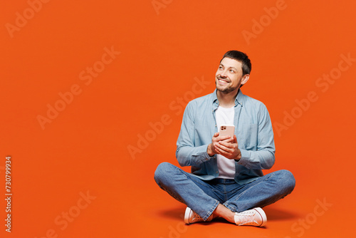 Full body young man he wearing blue shirt white t-shirt casual clothes sitting hold in hand use mobile cell phone look aside on area isolated on plain red orange background studio. Lifestyle concept. © ViDi Studio