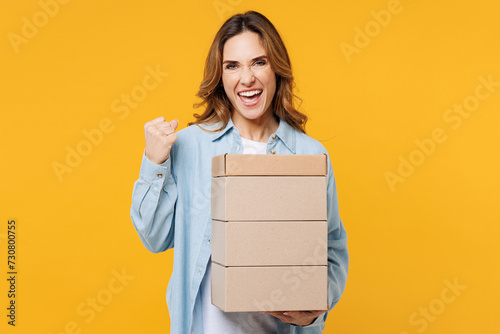 Overjoyed young happy woman she wear blue shirt white t-shirt casual clothes hold stack cardboard blank boxes do winner gesture isolated on plain yellow background studio portrait. Lifestyle concept. © ViDi Studio