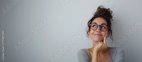 A happy, contemplative woman with glasses and a bun hairstyle gazing upwards, pondering, against a plain, light gray background with ample copy space. Suitable for Corporate Or Business Use