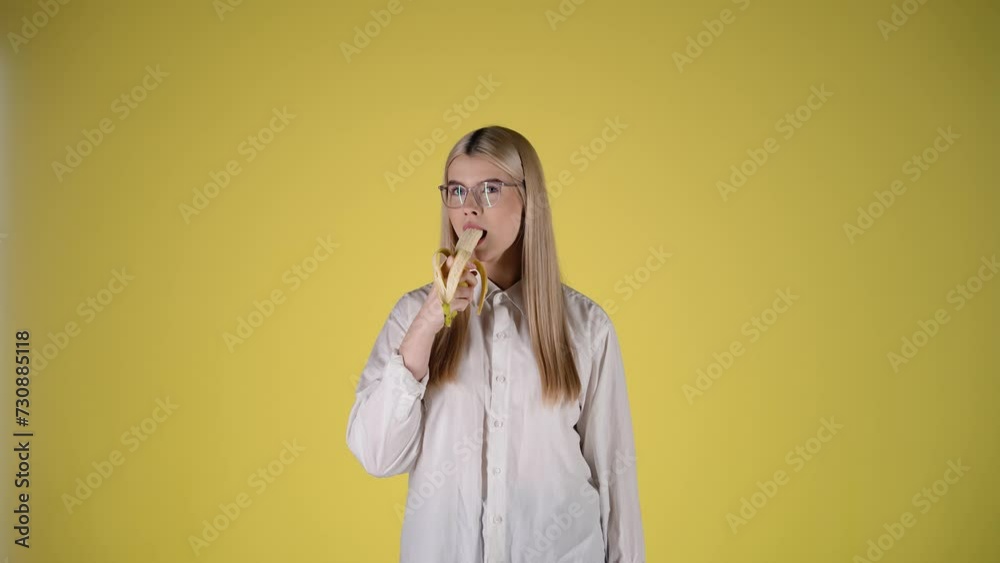 Blondynka w okularach i białej koszuli na żółtym tle gryzie banana i uśmiecha się prosto do kamery. Ujęcie lekkie, zabawne i sugestywne, z wyraźną ekspresją i kolorową estetyką
