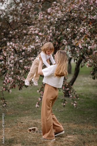 Beautiful mother and daughter against the background of a blooming apple tree. Mom raises her little daughter in her arms. Stylish clothes in neutral colors. Mom and daughter having fun