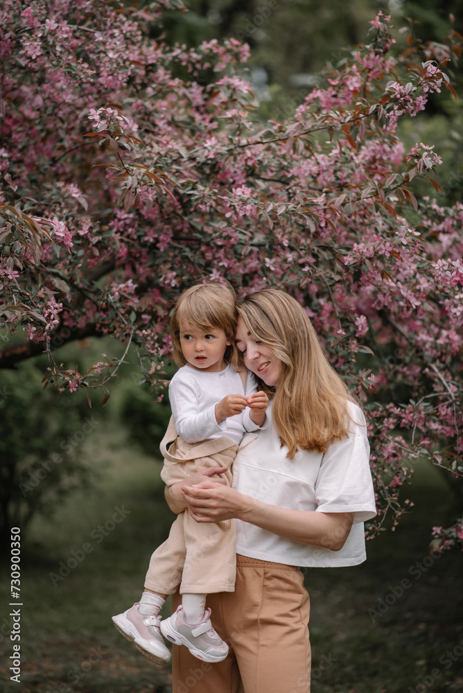 Beautiful mother and daughter against the background of a blooming apple tree. Mom holds her little daughter in her arms. Stylish clothes in neutral colors. Spring day in the park Beautiful mother and daughter against the background of a blooming apple tree. Mom holds her little daughter in her arms. Stylish clothes in neutral colors. Spring day in the park