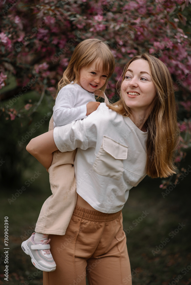 Beautiful mother and daughter against the background of a blooming apple tree. Mom rides her little daughter on her back. Stylish clothes in neutral colors. Mom and daughter having fun Beautiful mother and daughter against the background of a blooming apple tree. Mom rides her little daughter on her back. Stylish clothes in neutral colors. Mom and daughter having fun