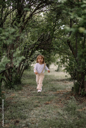 A small stylish girl  a child in beige trousers and a white blouse  runs in a tunnel from tall green bushes
