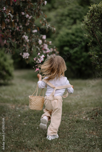 A little girl, a child in beige pants and a white blouse, with a basket in her hand, runs through the green grass with a twig in her hand. Green bushes of the park on a blurred background