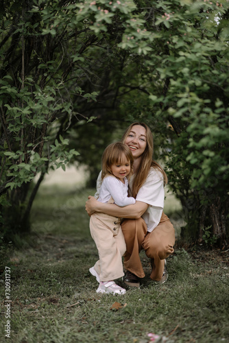 A small stylish girl, a child and her mother sat down on a green lawn in a park among bushes. Mom hugs her daughter. The daughter is holding a basket and a branch of a blossoming apple tree.