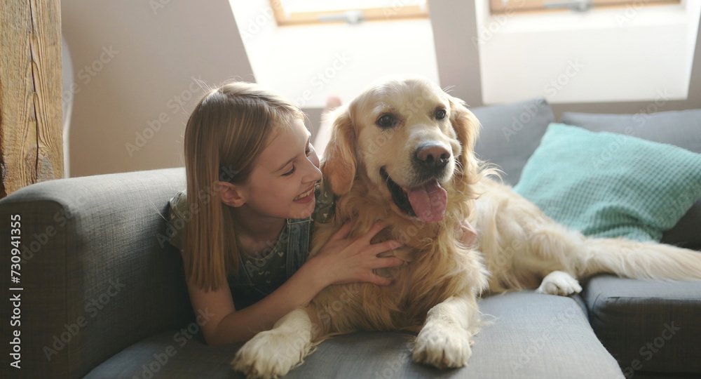 Child girl hugging golden retriever dog lying on sofa at home and smiling. Pretty preteen kid petting purebred pet doggy in loft room Child girl hugging golden retriever dog lying on sofa at home and smiling. Pretty preteen kid petting purebred pet doggy in loft room