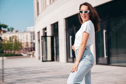 Young beautiful smiling hipster woman in trendy summer white t-shirt and jeans clothes. Sexy carefree model posing on the street background at sunset. Positive model outdoors. In sunglasses