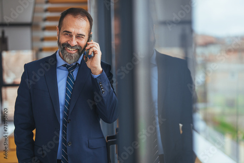 Chatting with pleasure. Portrait of happy mature qualified man is standing in light office and holding mobile phone. He is reading messages on gadget with smile. Copy space. 