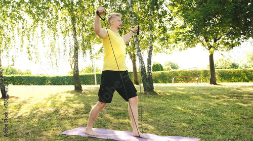 Athletic young man doing an exercises for the shoulders with the elastic rubber band on a grass Outdoors, healthy Sport activities and Fitness Concept