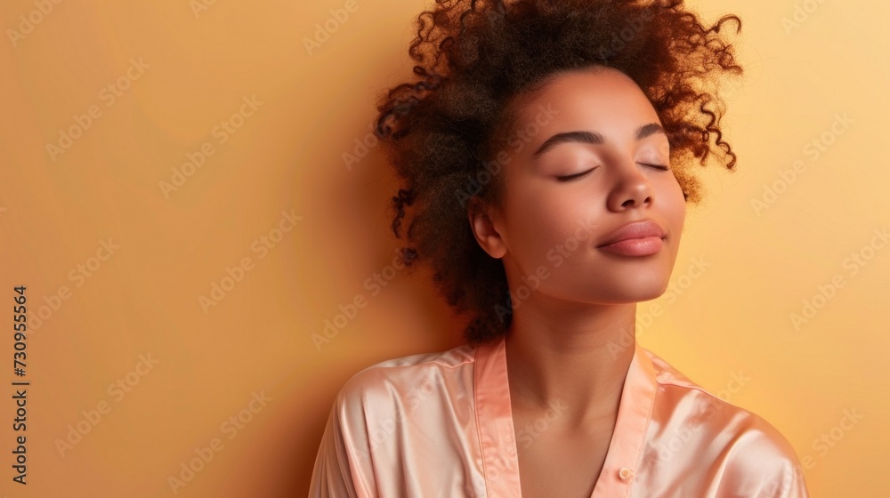 A young woman with curly hair wearing a pink blouse is captured in a serene moment with her eyes closed leaning against a warm orange background exuding a sense of tranquility and inner peace.
