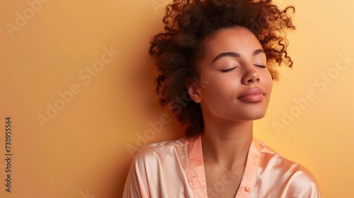 A young woman with curly hair wearing a pink blouse is captured in a serene moment with her eyes closed leaning against a warm orange background exuding a sense of tranquility and inner peace.
