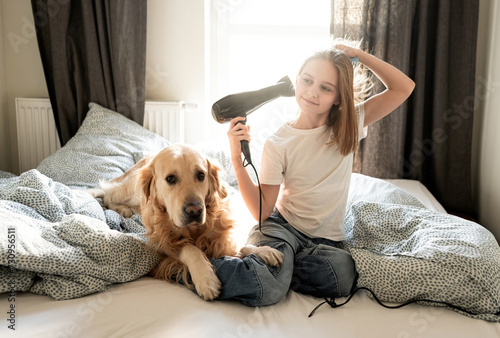Cute Little Girl Drying Hair With A Hairdryer, Sitting With Dog On A Bed