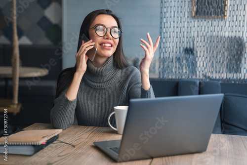 Happy business woman talking on cell phone sitting at table and working. Asian female sitting at office having telephonic conversation with client and smiling with look away photo
