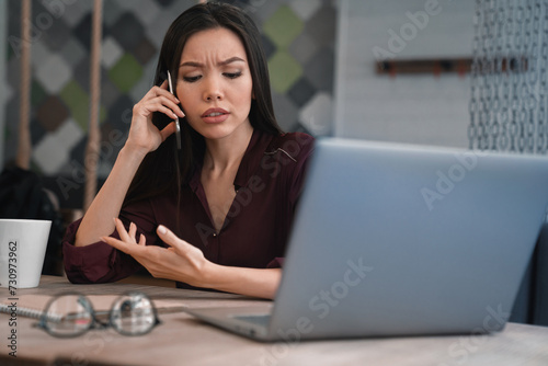 Busy woman sitting at her workplace and discussing with gesticulation some job questions on mobile phone in hand. Multitasking, deadline and workload