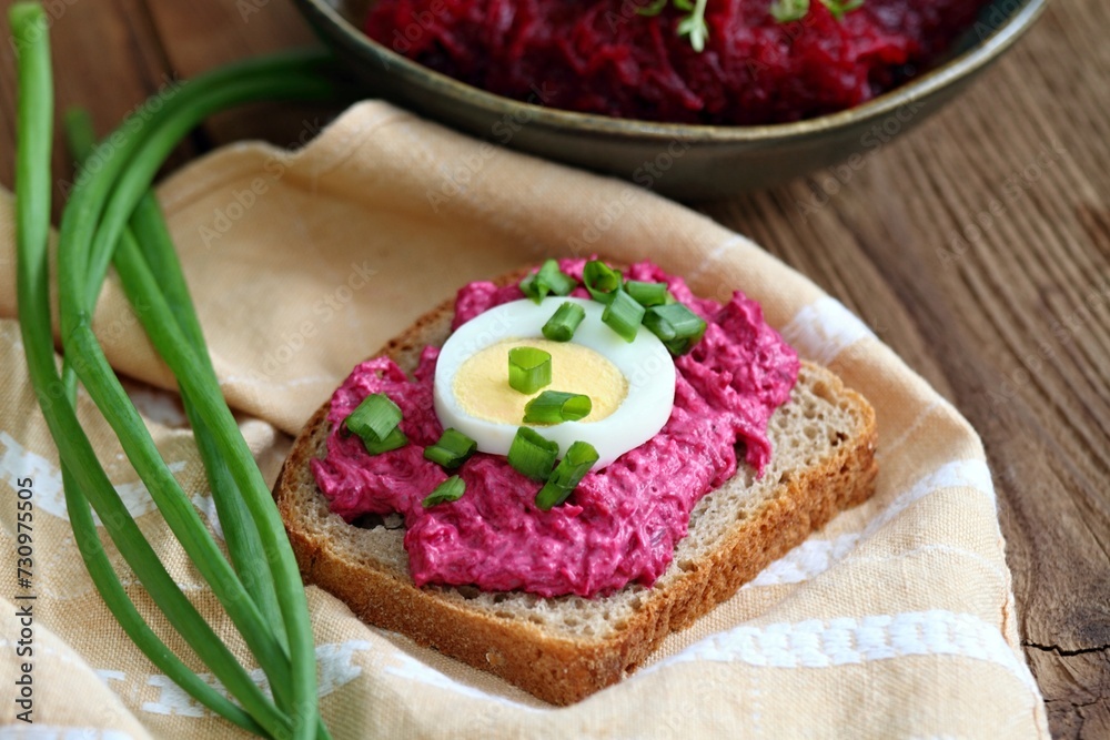 Pate spread from red beetroot on whole grain bread. Decorated with slice of egg and green onion. Traditional beetroot salad at back.