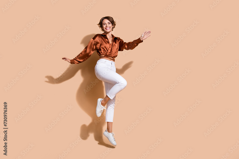 Full length photo of carefree shiny woman wear brown blouse jumping high empty space isolated beige color background