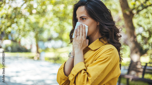 Portrait of a young brunette woman blowing her nose when standing close to flowers in a park. Woman with allergy symptoms blowing nose. © Dragana Gordic