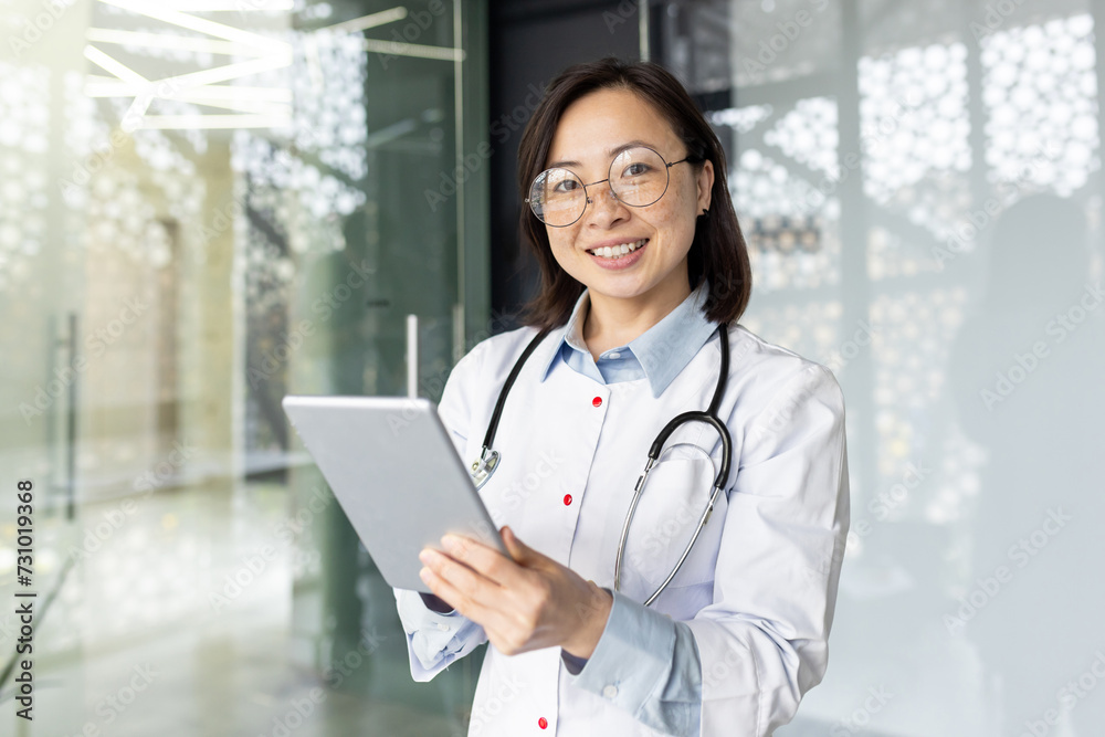 Smiling Asian female doctor with stethoscope using tablet for patient teleconsultation in a modern clinic office setting.