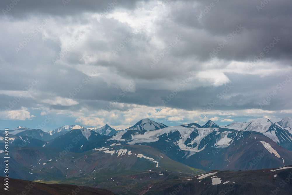 Scenic aerial view to green alpine valley, big rocky hills and large mountain range with snow-capped peaks in gray cloudy sky. Dramatic snowy mountain tops far away. Sunset light and grey rainy clouds