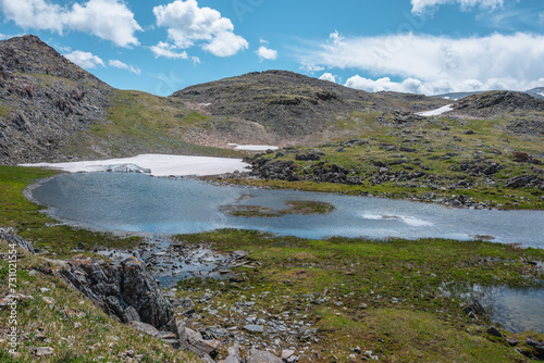 Beautiful small lake with snowfield among green grassy stony hills and rocks. Scenic view to alpine lake and rocky hilly mountains in sunlight under clouds in blue sky. Mountain lake in highlands.