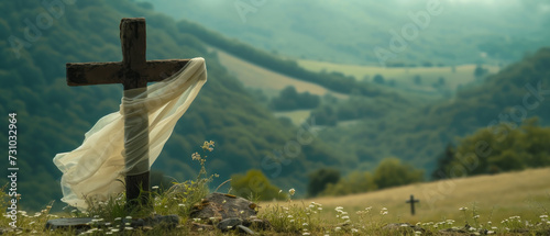 Sheer white scarf wrapped around wooden cross grave marker sitting on the edge of a precipice overlooking a verdant valley dotted with hay meadows and encircled by forested slopes. 