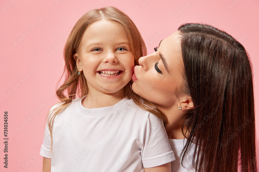 Caring, happy young woman, mother kissing her cute, smiling, tender little girl, daughter against pink studio background. Concept of happiness, Mother's day, childhood, fashion and lifestyle Caring, happy young woman, mother kissing her cute, smiling, tender little girl, daughter against pink studio background. Concept of happiness, Mother's day, childhood, fashion and lifestyle