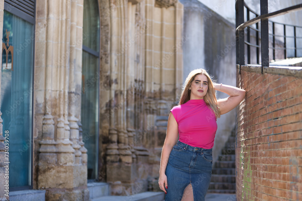Young woman, blonde, green eyes, wearing pink t-shirt and denim skirt, looking at camera, pensive and calm, leaning against a brick wall. Concept beauty, thoughts, dreams, tranquility.