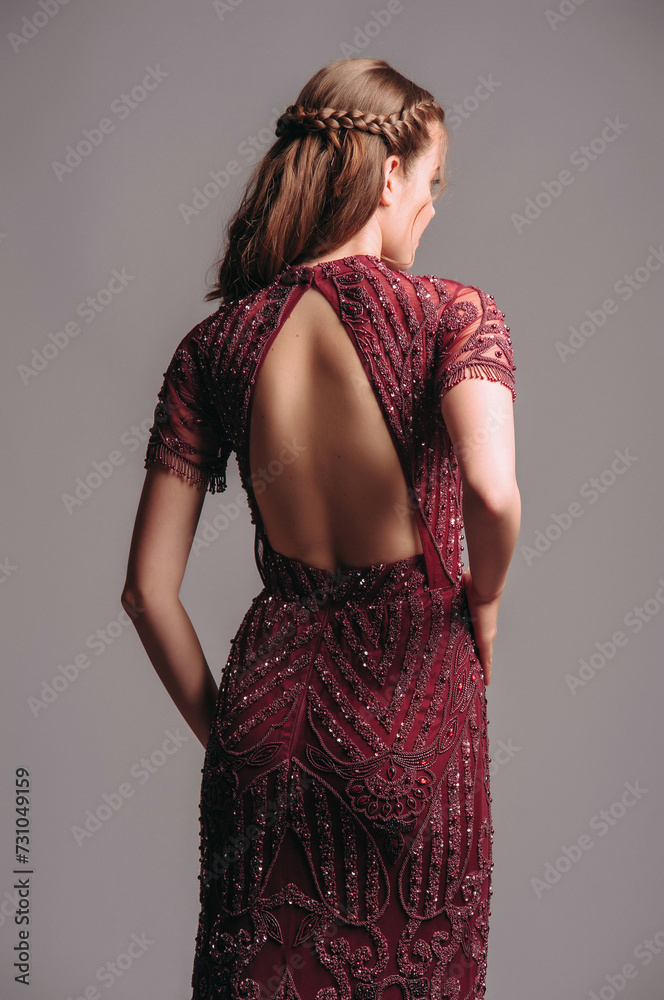 Fototapeta premium Bead embellished floral short sleeve maxi dress with back cut in maroon. Red backless transparent evening gown with a train. Beautiful young brunette woman's studio portrait. Female elegant clothes.
