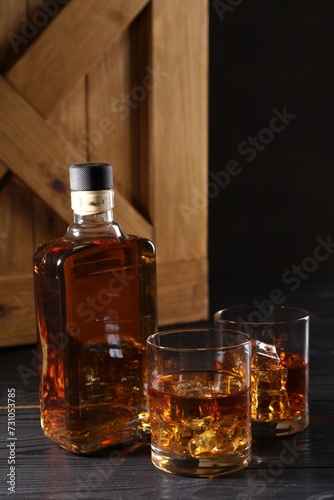 Whiskey with ice cubes in glasses and bottle on black wooden table